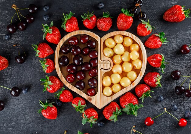 Cerezas con fresas, arándanos en un plato de madera sobre una superficie gris, plana.