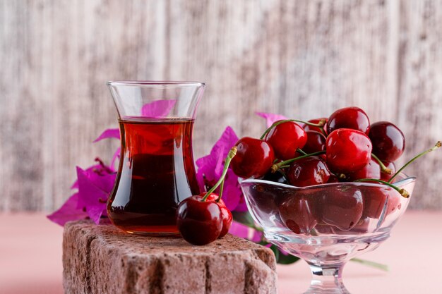 Cerezas en un jarrón con flores, vaso de té en vista lateral de ladrillo en superficie rosada y sucia