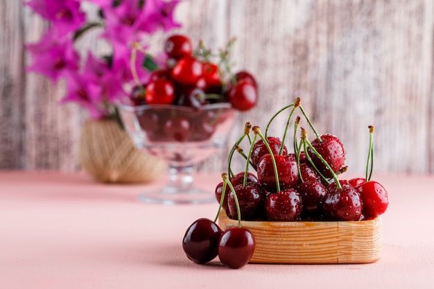 Cerezas con maceta en placa de madera y florero sobre superficie rosa y grunge, vista lateral