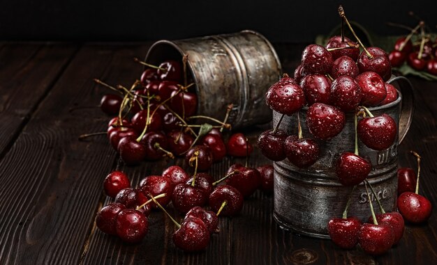 Cerezas maduras en gotas de agua, se encuentra en tazas de metal vintage. Fondo de madera oscura, enfoque selectivo. Cosecha fresca de cerezas jugosas