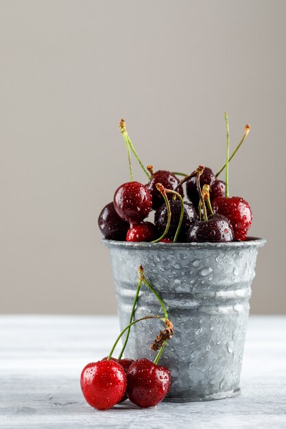 Cerezas en un mini cubo en grunge y superficie gris, vista lateral.