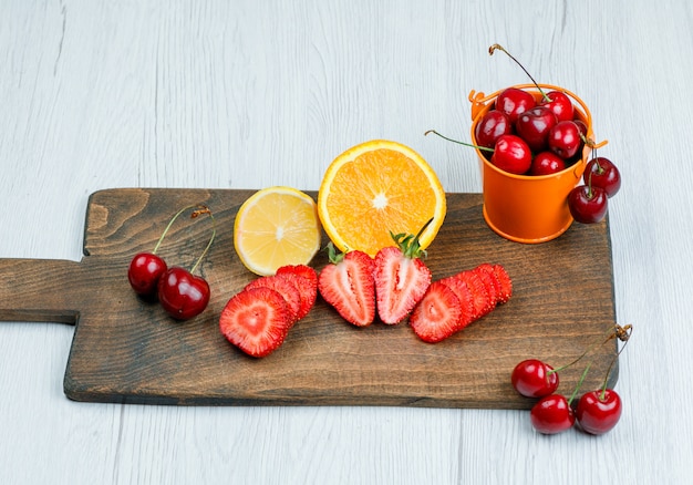 Cerezas en un mini cubo con limón, naranja, fresas planas sobre madera y tabla de cortar