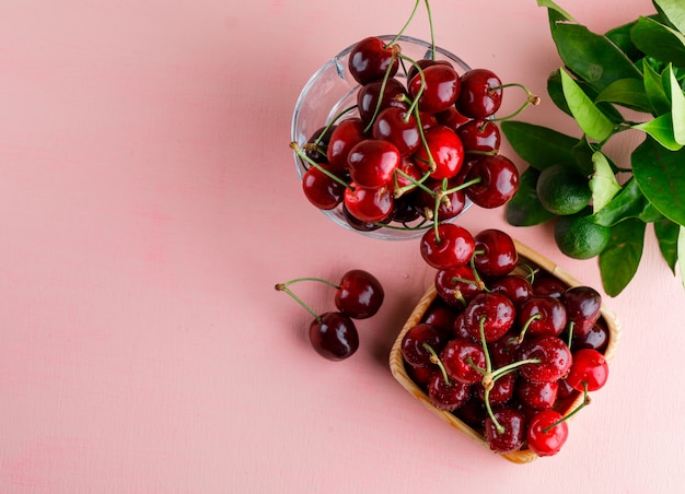 Cerezas en plato de madera y florero con limones y hojas vista superior sobre una superficie rosa