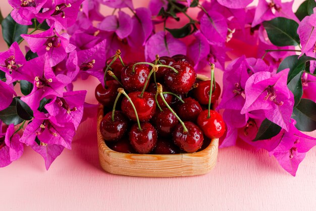 Cerezas en un plato de madera con flores vista de ángulo alto sobre una superficie rosa