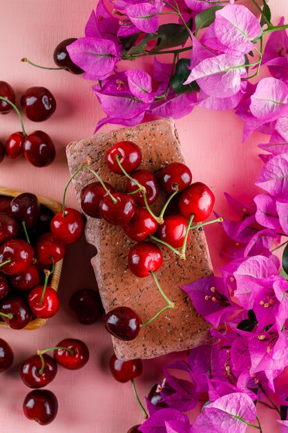 Cerezas en un plato de madera con flores, vista superior de ladrillo sobre una superficie rosa