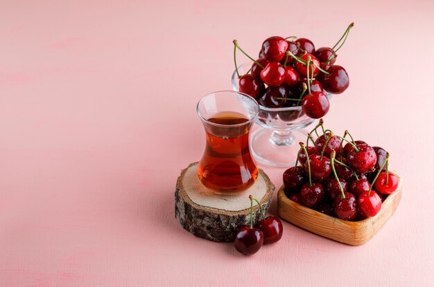 Cerezas con vaso de té sobre tabla de madera en placa de madera y florero sobre superficie rosa