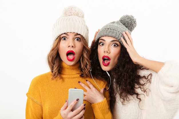 Cerrar chicas sorprendidas en suéteres y sombreros de pie junto con el teléfono inteligente sobre la pared blanca