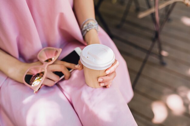 Cerrar detalles de manos de mujer sentada en la cafetería en traje de moda de verano, vestido de algodón rosa, gafas de sol, tomar café, accesorios elegantes, ropa relajante y moderna
