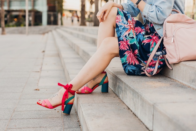 Cerrar detalles de piernas en sandalias rosas de mujer sentada en las escaleras en la calle de la ciudad con elegante falda estampada con mochila de cuero, tendencia de estilo veraniego