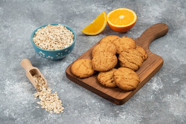 Cerrar foto de galletas caseras sobre tabla de madera y avena con naranjas sobre mesa gris.