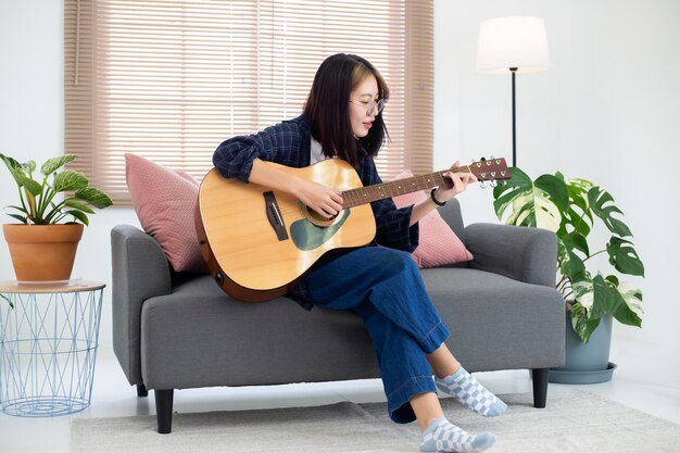 Cerrar gafas felices Chica asiática tocando la guitarra acústica en la sala de estar en casa Concepto de recreación en casa