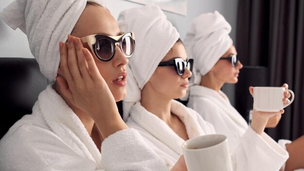 Cerrar hermosas chicas en albornoces blancos con toallas en la cabeza y gafas de sol tomando café en la cama en la habitación del hotel