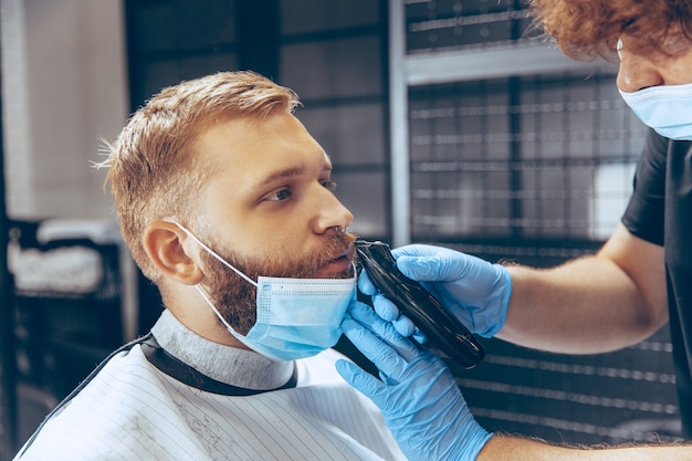 Cerrar hombre cortándose el pelo en la barbería con máscara durante la pandemia de coronavirus.