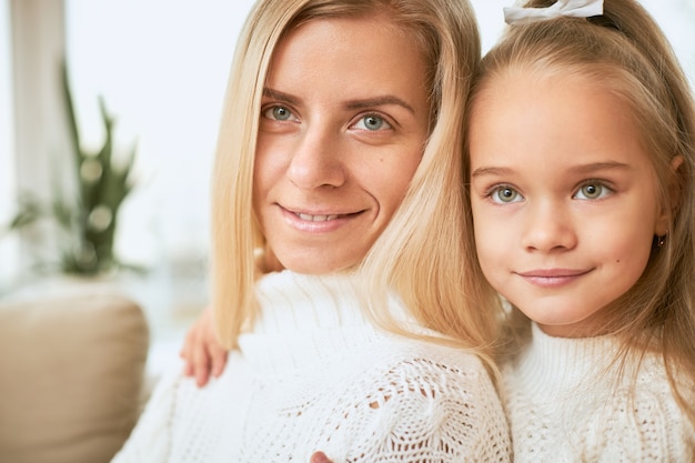 Cerrar imagen de alegre linda niña sentada detrás de feliz joven madre abrazándola fuerte, disfrutando de un buen rato juntos en casa. Concepto de personas, infancia, familia, vinculación y relaciones.