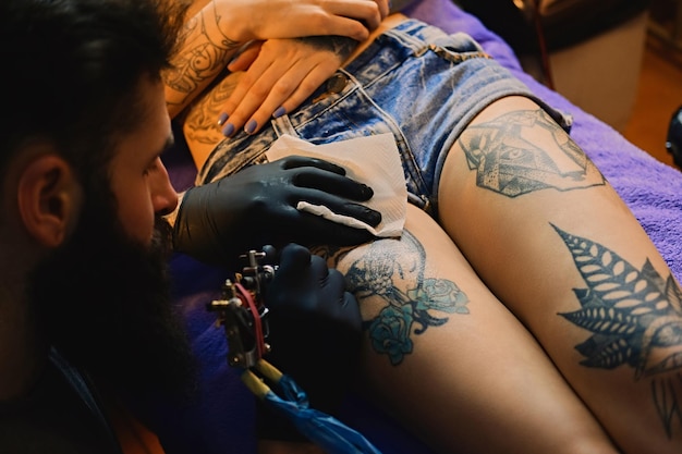 Cerrar la imagen del artista masculino del tatuaje barbudo hace un tatuaje en una pierna femenina.
