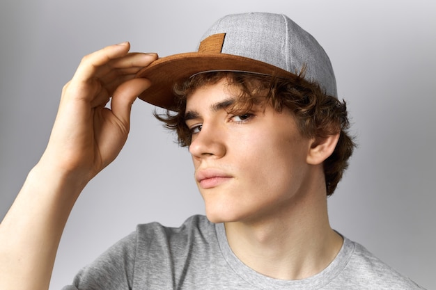 Cerrar imagen de chico europeo joven guapo con pelo rizado y cara suave divirtiéndose vistiendo snapback moderno sosteniendo la mano en el pico y mirando a la cámara con mirada segura Estilo y vestimenta