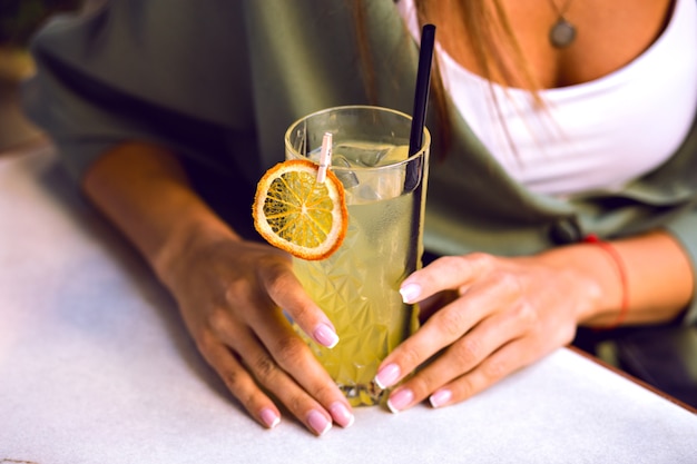 Cerrar imagen de detalles de mujer sosteniendo cóctel de limonada sabroso fresco, hermosas manos con manicura francesa, ropa casual elegante, colores tonificados.