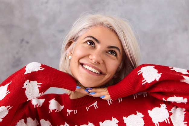 Foto gratuita cerrar imagen de encantadora joven mujer europea de moda en elegante jersey de punto rojo soñando o pensando en algo agradable, mirando hacia arriba con una radiante sonrisa dentuda, tomados de la mano debajo de la barbilla