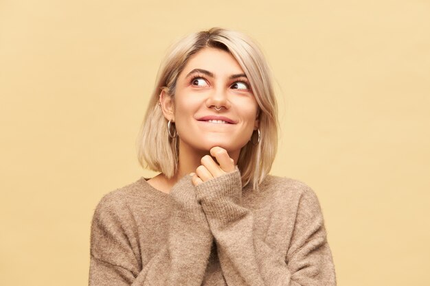 Cerrar imagen de encantadora joven mujer rubia con anillo en la nariz y corte de pelo bob tomados de la mano debajo de la barbilla y mirando a otro lado con una sonrisa misteriosa y juguetona, jugando bromas y haciendo travesuras