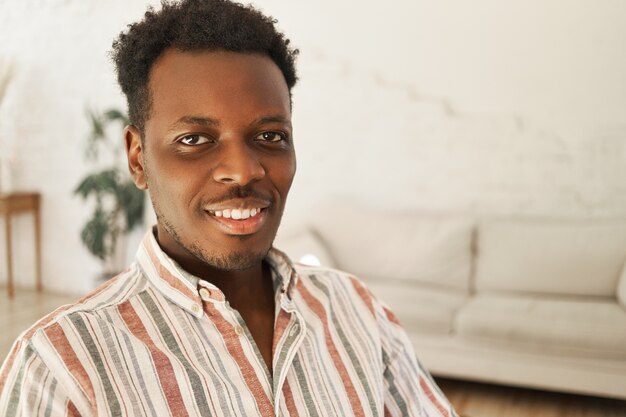 Cerrar imagen de hombre de piel oscura joven alegre fresco con peinado afro sentado en el elegante salón interior, relajarse en casa, mirando a cámara con amplia sonrisa feliz.