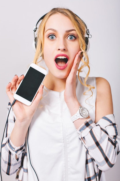 Cerrar imagen interior de mujer linda joven alegre escuchando música con auriculares, sosteniendo el teléfono móvil, mirando con ojos sorprendidos, tocando la cara. El uso de ropa casual con estilo.