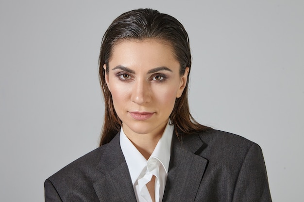 Cerrar imagen de mujer andrógina de aspecto moderno con maquillaje y cabello peinado hacia atrás posando, vestida con una chaqueta de hombre gris de gran tamaño, mirando con expresión facial seria