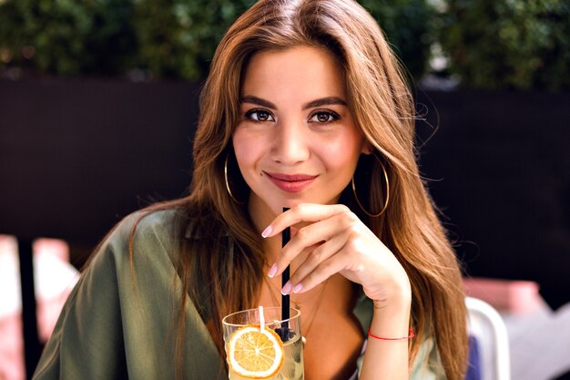 Cerrar imagen de mujer bonita morena bebiendo limonada sabrosa y mirando a cámara, disfrutando del tiempo en la terraza de la ciudad, verano cálido, belleza natural.