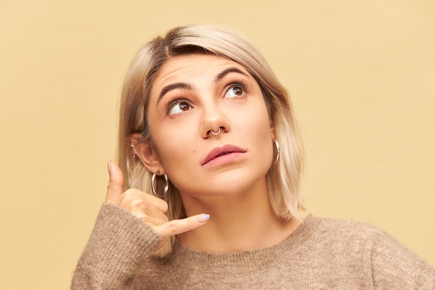 Cerrar imagen de mujer bonita con peinado bob rubio y anillo en la nariz mirando hacia arriba, sosteniendo la mano en la oreja con el pulgar y el meñique extendidos ampliamente, haciendo el gesto Llámame. Lenguaje corporal