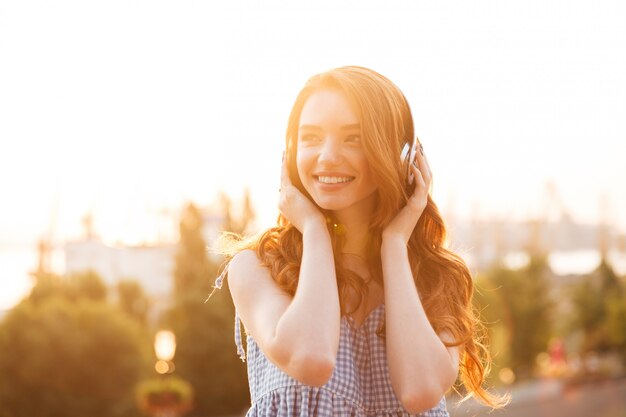 Cerrar imagen de mujer joven belleza jengibre en vestido escuchando música en la puesta de sol