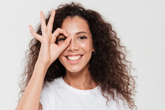 Cerrar imagen de mujer rizada sonriente mano en la cara y mostrando signo ok