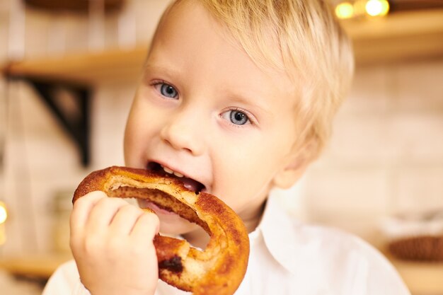 Cerrar imagen recortada de bebé caucásico con cabello rubio y ojos azules abriendo la boca para morder bagel crujiente, con expresión facial alegre. Concepto de infancia, alimentación, cuidado y salud.