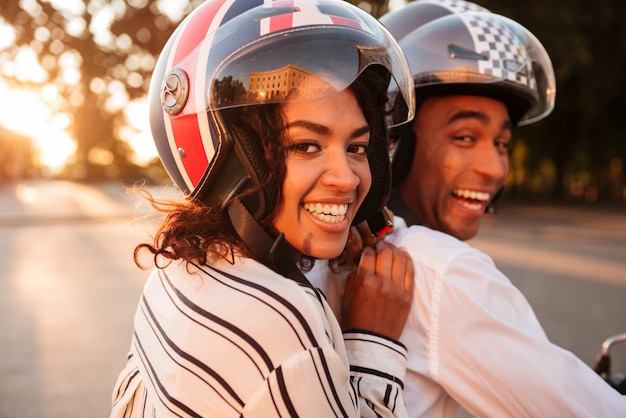 Cerrar imagen de vista lateral de la feliz pareja africana monta en moto moderna al aire libre y mirando a la cámara