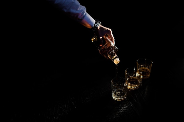Cerrar la mano sosteniendo la botella vierte la bebida en un vaso en un espacio oscuro negro Concepto de celebración y cócteles de barman de despedida de soltero