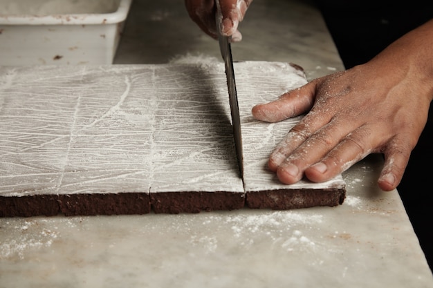 Cerrar las manos del hombre negro cortar una rebanada de pastel de chocolate recién horneado sobre una mesa de mármol en confitería artesanal profesional