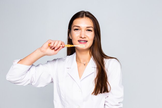 Cerrar mujer médico sonriendo con brackets de cerámica y metal con cepillo de dientes para cepillar los dientes en la pared blanca, belleza y concepto saludable