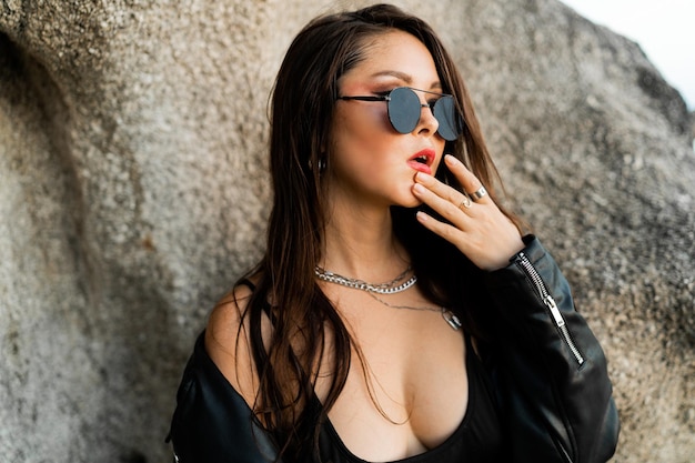 Foto gratuita cerrar op retrato de mujer confiada con labios rojos en chaqueta de cuero negro de moda y sensual vestido negro posando sobre fondo rocoso