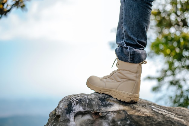 Cerrar la pierna del hombre de senderismo en jeans con zapatillas de trekking colocadas en el pico de la montaña rocosa en el cielo azul del bosque natural en el espacio de copia de fondo