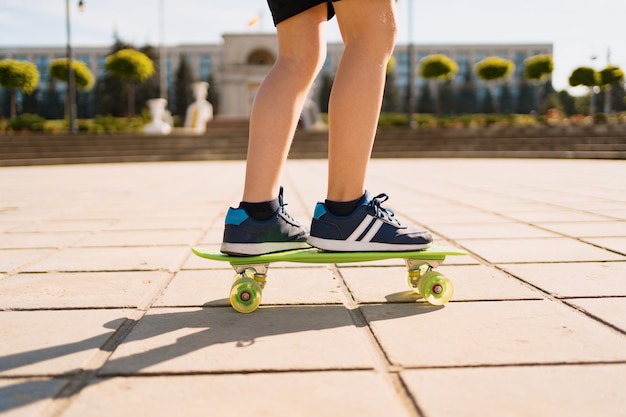 Cerrar las piernas en zapatillas azules montando en patineta verde en movimiento. Estilo de vida urbano activo de juventud, formación, hobby, actividad. Deporte activo al aire libre para niños. Niño en patineta.