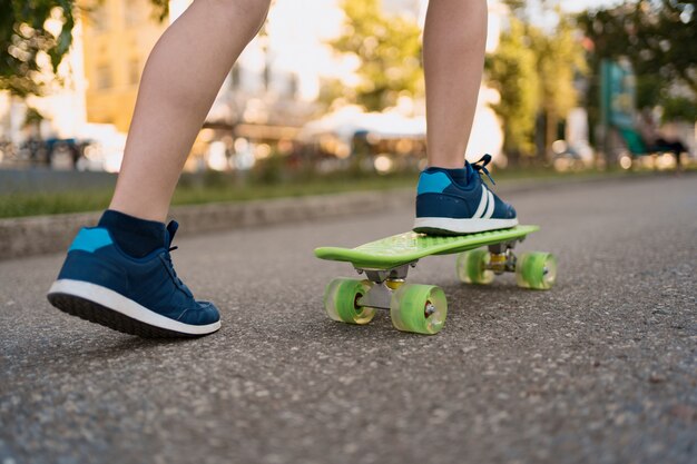 Cerrar las piernas en zapatillas azules montando en patineta verde en movimiento. Estilo de vida urbano activo de juventud, formación, hobby, actividad. Deporte activo al aire libre para niños. Niño en patineta.