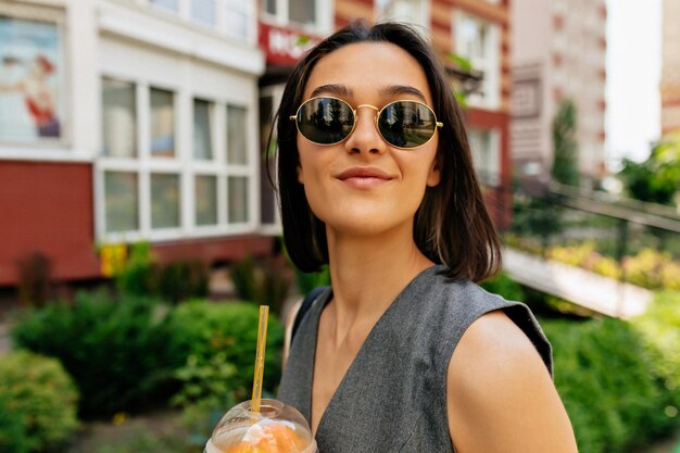 Cerrar el retrato al aire libre de una mujer muy sonriente con el pelo corto y oscuro con gafas de sol sosteniendo una bebida de verano sonriendo a la cámara en el fondo de la ciudad