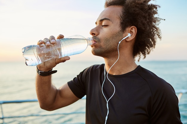 Cerrar retrato de atleta afroamericano en forma bebiendo agua de una botella de plástico con auriculares. Refrescándose con agua y vistiendo camiseta negra