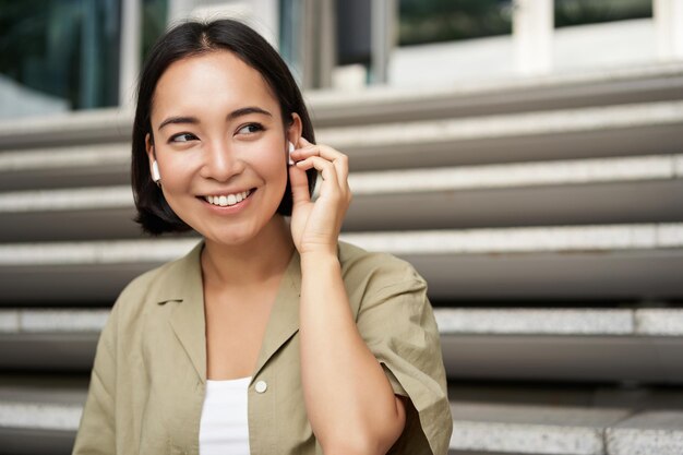 Cerrar retrato calle foto de mujer asiática escuchando música en auriculares inalámbricos sonriendo sentado