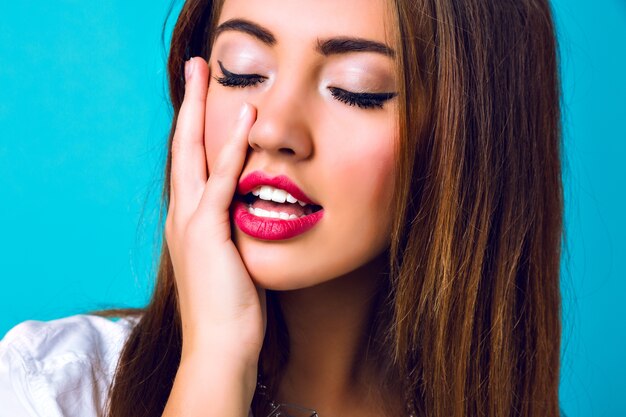 Cerrar retrato de chica de moda elegante, piel perfecta, maquillaje brillante, pelos morenos, pura belleza, cerrar los ojos y sonreír, dientes blancos y labios carnosos, fondo de estudio azul.