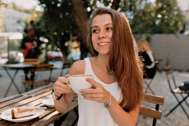 Cerrar retrato de hermosa niña caucásica con cabello largo sentado en la terraza de verano de espacio abierto con una taza de café