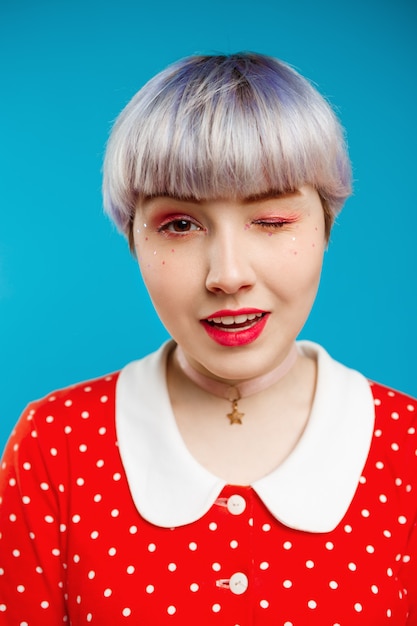 Foto gratuita cerrar retrato hermosa niña dollish con corto cabello violeta claro con vestido rojo guiñando un ojo sobre la pared azul