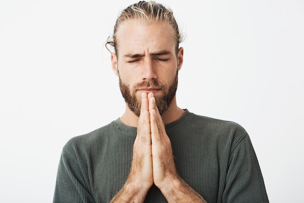 Cerrar retrato de hombre maduro concentrado con elegante corte de pelo y barba cogidos de la mano juntos delante de la cara, rezando y esperando
