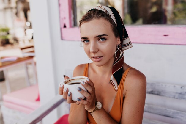 Cerrar retrato de mujer adorable con accesorios en la cabeza sosteniendo café y mirando mientras desayuna afuera