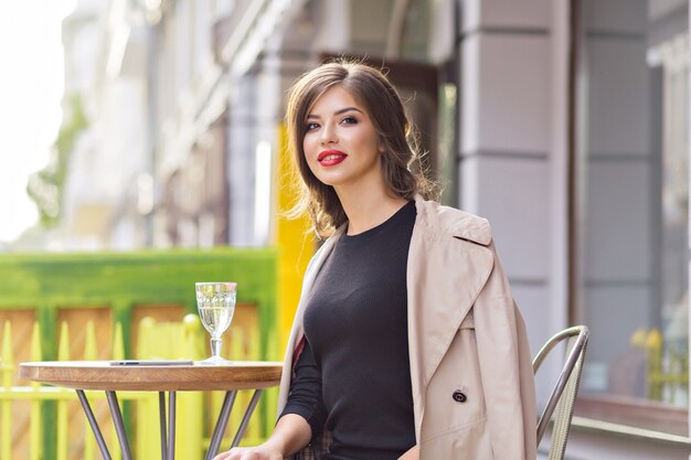 Cerrar retrato de mujer bonita encantadora con labios rojos con vestido negro y abrigo beige descansando en la cafetería de verano con un vaso de agua