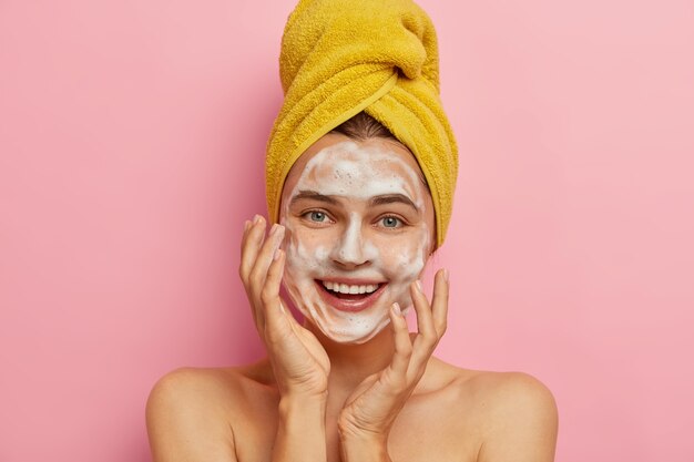 Cerrar retrato de mujer caucásica feliz que se lava la cara con agua y jabón facial, quiere tener una tez saludable, elimina la suciedad y el sebo del sudor, toalla envuelta en amarillo en la cabeza