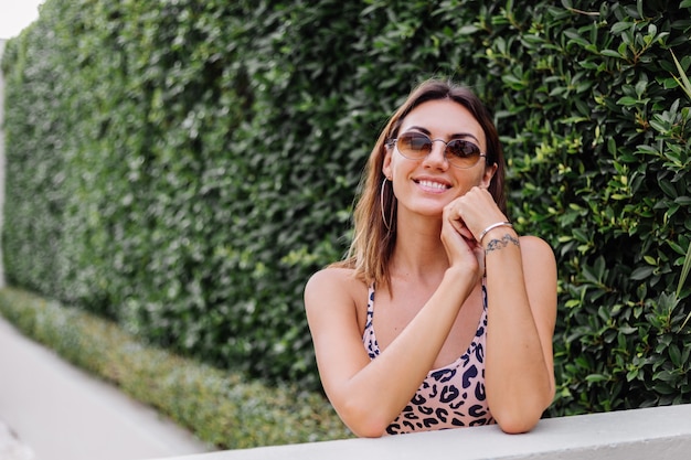 Cerrar retrato de mujer europea en top de leopardo y gafas de sol y aretes redondos en la pared de arbusto verde